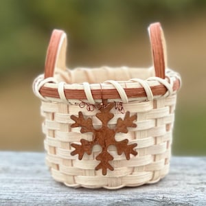 May include: A small woven basket featuring light beige reed construction with two upright handles. The rim is accented with a darker brown band, and a metallic, rust-colored snowflake ornament is attached to the front with thin wire.