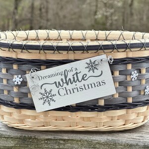 May include: Woven basket with a rectangular shape, featuring a light beige base, gray and black accents, and a decorative tag. The tag reads "Dreaming of a white Christmas" with snowflake designs. The basket is adorned with snowflake embellishments.