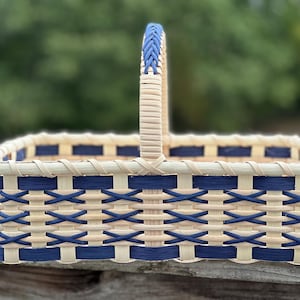 May include: A rectangular woven basket featuring a light beige natural reed base with alternating navy blue horizontal and vertical strips. The basket includes a single upright handle with a braided navy blue accent at the top centre.