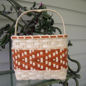 May include: A handcrafted basket with a handle, featuring a woven design in natural and reddish-brown tones. The basket's rectangular body has a decorative pattern, and the handle is arched. The basket is displayed on a glass surface.
