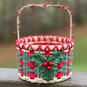 May include: A small woven basket with a red and green plaid pattern and a holly design. The basket has a handle made of twigs with red berries.