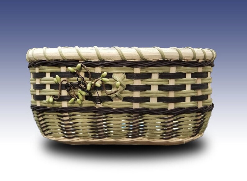 May include: A woven basket with a green and brown pattern. The basket has a decorative vine with green leaves and berries.