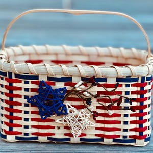 May include: A rectangular woven basket with a handle, featuring a patriotic red, white, and blue color scheme. The basket is adorned with star-shaped decorations and sprigs of berries, perfect for decorative use.