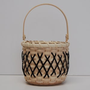 May include: A small, woven basket with a black and white diamond pattern. The basket has a wooden handle and is made of natural materials.