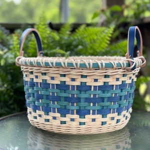 May include: A woven basket with blue and green accents and leather handles. The basket is made of natural materials and has a rustic look.