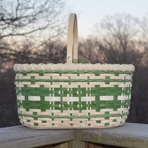 May include: A green and natural woven basket with a wooden handle. The basket is oval shaped and has a checkered pattern.