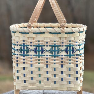 May include: A rectangular woven basket with a wooden handle and legs. The basket features a cream-colored base with blue and teal accents in a geometric pattern. The handle and legs are made of light-colored wood.