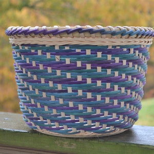 May include: A woven basket with a light brown base and purple and teal colored woven strips. The basket is sitting on a wooden surface.