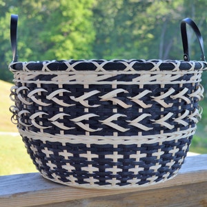 May include: A black and white woven basket with two black leather handles. The basket is made of natural materials and has a unique pattern.