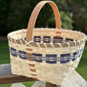 Puede incluir: Una cesta tejida con un asa de madera natural y un patrón de cuadros azul y gris. La cesta está hecha de materiales naturales y tiene un aspecto rústico.