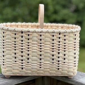 May include: A rectangular woven basket featuring a light beige natural finish with vertical dark brown accent strips. The basket includes a single upright handle, a braided top rim, and small rounded feet at the base for stability.