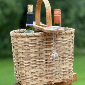 Puede incluir: Una cesta de vino de mimbre tejido con un asa de madera y un soporte para una copa de vino. La cesta contiene dos botellas de vino, una con corcho y otra con tapón naranja. La cesta está sobre una superficie de madera.