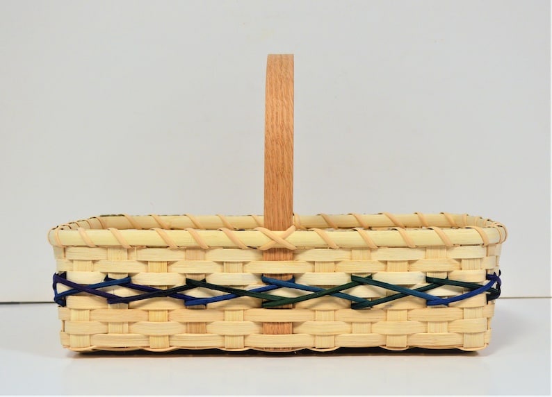 Op de afbeelding: Een rechthoekige geweven mand met een handvat van natuurlijk hout. De mand heeft een decoratieve geweven rand in blauw, groen en wit.