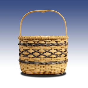 May include: A small, round woven basket with a handle. The basket is made of light brown wicker with a decorative dark brown and black woven pattern around the middle. The handle is arched and attached to the top of the basket.