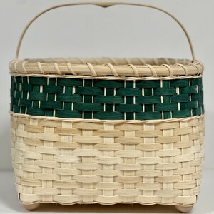 May include: A woven basket with a green band and a natural wood handle. The basket has a square shape and is made of natural materials.