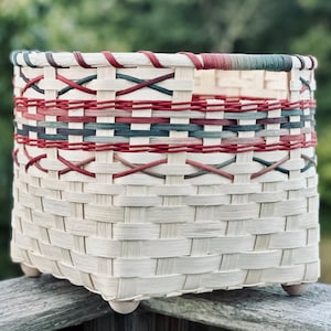 May include: A woven basket featuring a light cream base with horizontal bands of red and dark green accents. The basket has a square-shaped body with a flat rim and four small, rounded wooden feet attached to the bottom corners for stability.