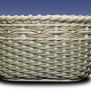May include: A woven basket with a light green and natural colour pattern. The basket is oval shaped and has a woven rim.