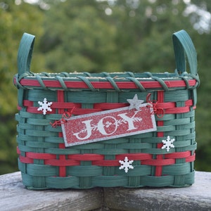 May include: A rectangular woven basket featuring dark green and red splints with two upright handles. The front is decorated with three white snowflake ornaments and a rectangular red glitter sign with the white text JOY.