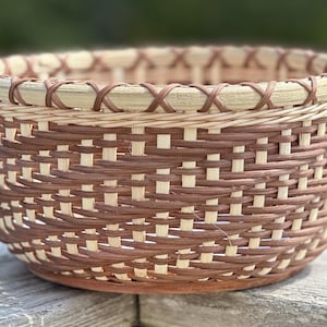 May include: A round, handwoven basket with a natural and brown color scheme. The basket features a woven pattern with a decorative rim. The base is a darker brown, and the rim has a decorative cross-stitch design.