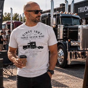 May include: A light grey short-sleeved t-shirt featuring a black graphic of a semi-truck and text that reads SINCE 1987 THREE SEVEN NINE ASSEMBLY DENTON TEXAS MODEL 379 THE DESIGN OUTLAW YOUR PURVEYOR OF FINELY CRAFTED GOODS.