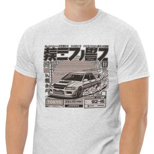 Puede incluir: Camiseta gris claro con un gráfico en blanco y negro de un coche derrapando. El gráfico incluye texto japonés, la palabra "TOKYO" y los números "92-16". La camiseta está hecha de un material suave y cómodo.