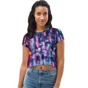 Può includere: Una maglietta corta con un design di paesaggio urbano futuristico. L'opera presenta insegne al neon nei toni del rosa, blu e viola, con una figura solitaria in lontananza. La maglietta è realizzata in un materiale morbido e confortevole.