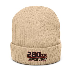 Puede incluir: Un gorro de punto acanalado beige con el texto bordado "280zx SINCE 1978" en negro y rojo. El gorro está doblado en el borde y tiene un estilo clásico e informal.