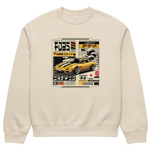 May include: A cream-coloured sweatshirt with a graphic of a yellow sports car, Japanese text, and a wave design. The car is set against a backdrop of Japanese-inspired elements, including red and black accents. The sweatshirt has a crew neck and long sleeves.