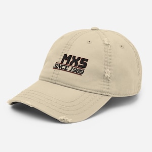 Puede incluir: Una gorra de béisbol desgastada de color beige con texto bordado en negro y rojo que dice "MX5 SINCE 1989".