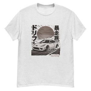 May include: Light gray t-shirt featuring a graphic of a white sports car drifting on a road. The design includes Japanese text and a sun graphic. The text "DESIGN OUTLAW" is at the bottom of the graphic.