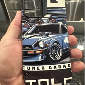 May include: A black and white phone case with a blue and white illustration of a vintage car. The car has the number "440" on the side and the text "TUNER GARAGE" below it. The case also has the text "TOLC" on the bottom.