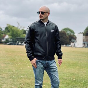 May include: A black bomber jacket with a white logo is worn with blue jeans. The jacket has a zip closure and ribbed cuffs and hem. The person is wearing sunglasses and standing on a grassy field.