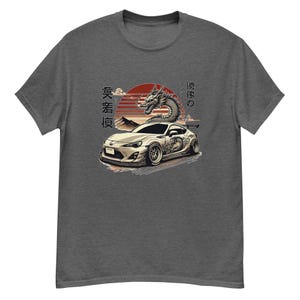 以下が含まれることがあります： ヘザーグレーのTシャツに、白い車のグラフィックが描かれています。車にはドラゴンがデザインされています。車は夕焼けをバックに、日本語で「藤原」「フライング」「真香寿」とプリントされています。