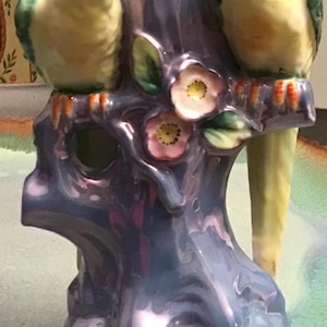 May include: A ceramic parrot figurine perched on a tree trunk base. The parrots have yellow and green plumage with orange accents. The base is a glossy, iridescent blue with pink flowers.