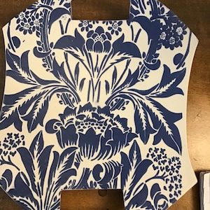 May include: A decorative, off-white ceramic tile with a blue floral pattern. The design features stylized flowers and leaves, with a handle at the top and a cutout at the bottom. The tile is approximately 10 inches wide and 12 inches tall.