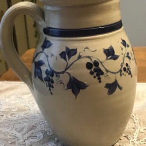 May include: A beige ceramic pitcher with a curved handle and a spout. The pitcher is decorated with blue grape vines and a dark blue band around the neck. The pitcher sits on a white lace tablecloth.