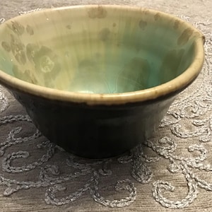 May include: A handmade ceramic bowl with a unique glaze. The bowl features a dark base that transitions to a light green interior with a speckled pattern. The rim is a warm, earthy brown. This decorative bowl is ideal for serving or display.