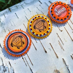 May include: Three circular pins featuring orange centres with the text Every Child Matters. The pins are decorated with intricate beadwork in orange, black, white, yellow, and red, arranged in concentric patterns around the central printed design.