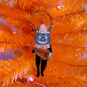 May include: A black cat ornament featuring a paper cutout face with a wide grin, wearing a peach-colored party hat with blue polka dots and a matching ruffled collar. The cat has a gold tinsel halo and holds a small, brown bottle brush tree.