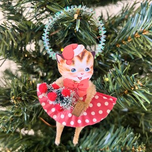 May include: A vintage-style Christmas ornament featuring a cartoon cat wearing a Santa hat and a red polka-dot dress. The cat holds a bouquet of red pom-poms and greenery. A sparkly halo encircles the cat's head, creating a festive holiday decoration.