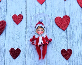 May include: A vintage-style Valentine's Day ornament featuring a cherubic figure in a red outfit and hat, surrounded by red glitter hearts. The ornament has a halo and holds heart-shaped bells. The background is a light blue wooden surface.
