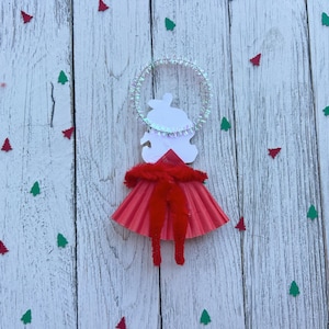 May include: A handmade Christmas ornament in the shape of an angel. The angel has a white paper body, a red skirt, and a halo. The ornament is decorated with red and green tree-shaped confetti on a white wooden background.