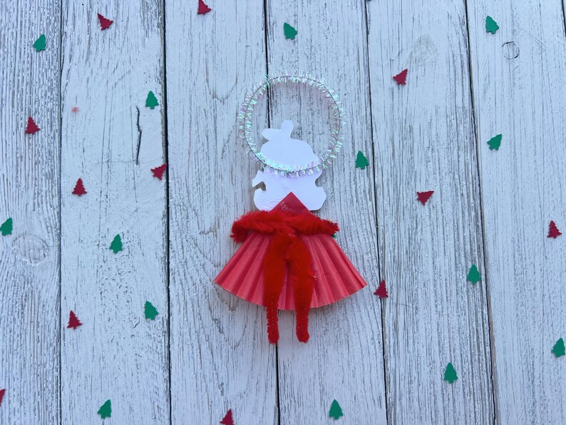 May include: A handmade Christmas ornament in the shape of an angel. The angel has a white paper body, a red skirt, and a halo. The ornament is decorated with red and green tree-shaped confetti on a white wooden background.
