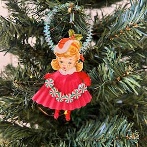 May include: A vintage Christmas tree ornament featuring a girl in a red dress and hat, adorned with candy cane accents. The ornament is suspended from a shimmering, iridescent garland, set against a backdrop of green Christmas tree branches.