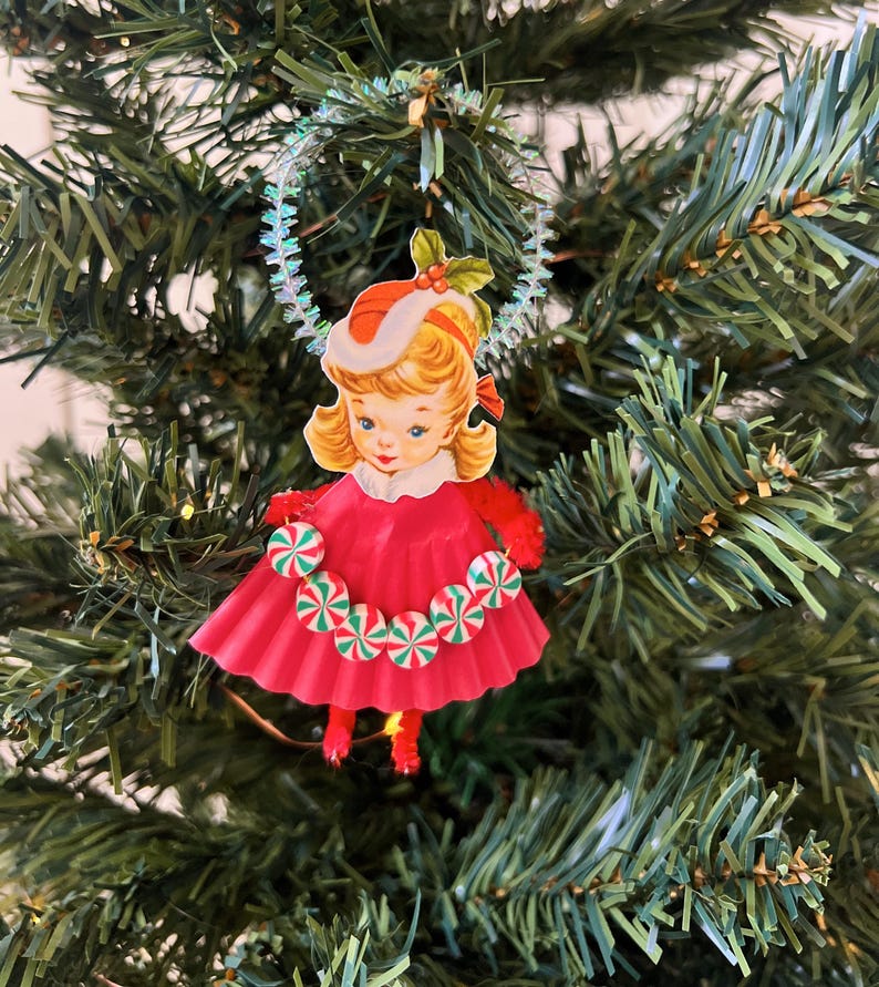 May include: A vintage Christmas tree ornament featuring a girl in a red dress and hat, adorned with candy cane accents. The ornament is suspended from a shimmering, iridescent garland, set against a backdrop of green Christmas tree branches.
