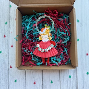May include: A vintage-style Christmas ornament of a girl in a red dress and hat, surrounded by red and teal paper shreds inside a cardboard box. The ornament has a white and green garland around the neck. The box sits on a white wooden surface.