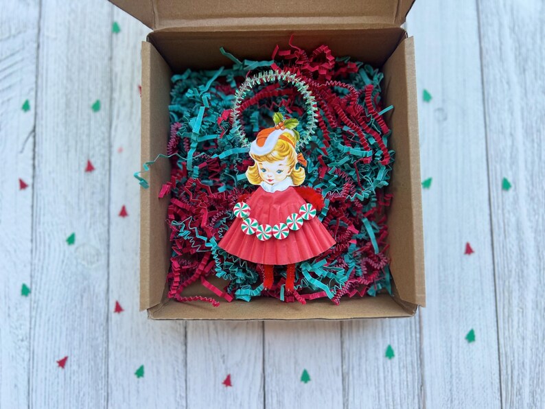 May include: A vintage-style Christmas ornament of a girl in a red dress and hat, surrounded by red and teal paper shreds inside a cardboard box. The ornament has a white and green garland around the neck. The box sits on a white wooden surface.