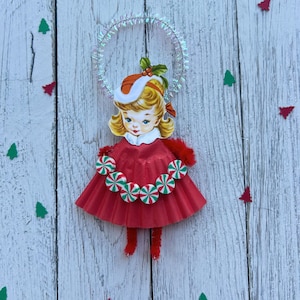 May include: A vintage-style Christmas ornament featuring a girl in a red dress adorned with candy-shaped embellishments. The ornament has a halo and is set against a white wooden background with scattered Christmas tree and triangle cutouts.