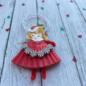 May include: A vintage Christmas ornament in the shape of a girl wearing a red dress and a Santa hat. The ornament has a halo and a candy cane necklace. Green and red tree confetti is scattered around the ornament.
