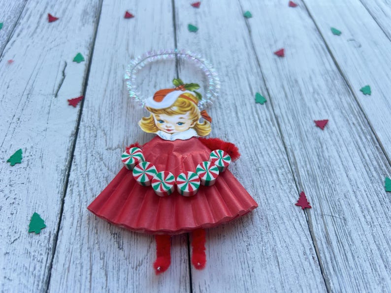 May include: A vintage Christmas ornament in the shape of a girl wearing a red dress and a Santa hat. The ornament has a halo and a candy cane necklace. Green and red tree confetti is scattered around the ornament.